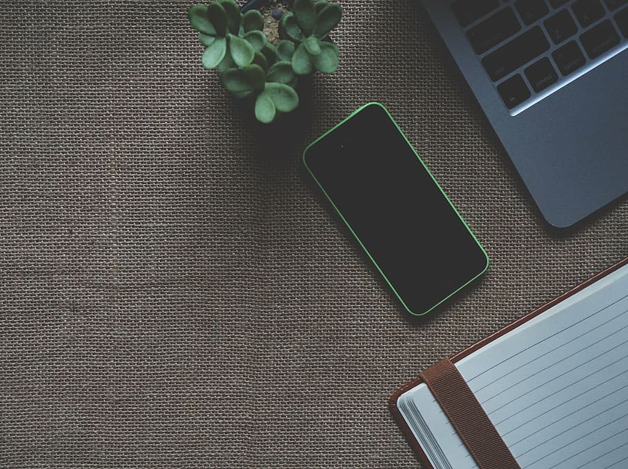Overhead view of a smartphone, a laptop, a notebook, and a small plant on a textured surface.